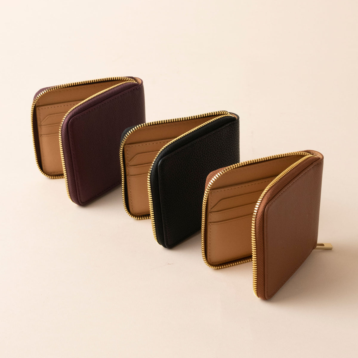 Three small leather zip wallets in burgundy, black, and brown arranged in a row with tan interiors and gold zippers.