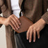 A person places a caramel brown rectangular zip wallet into a black bag. The wallet has a gold zipper and pebbled texture.