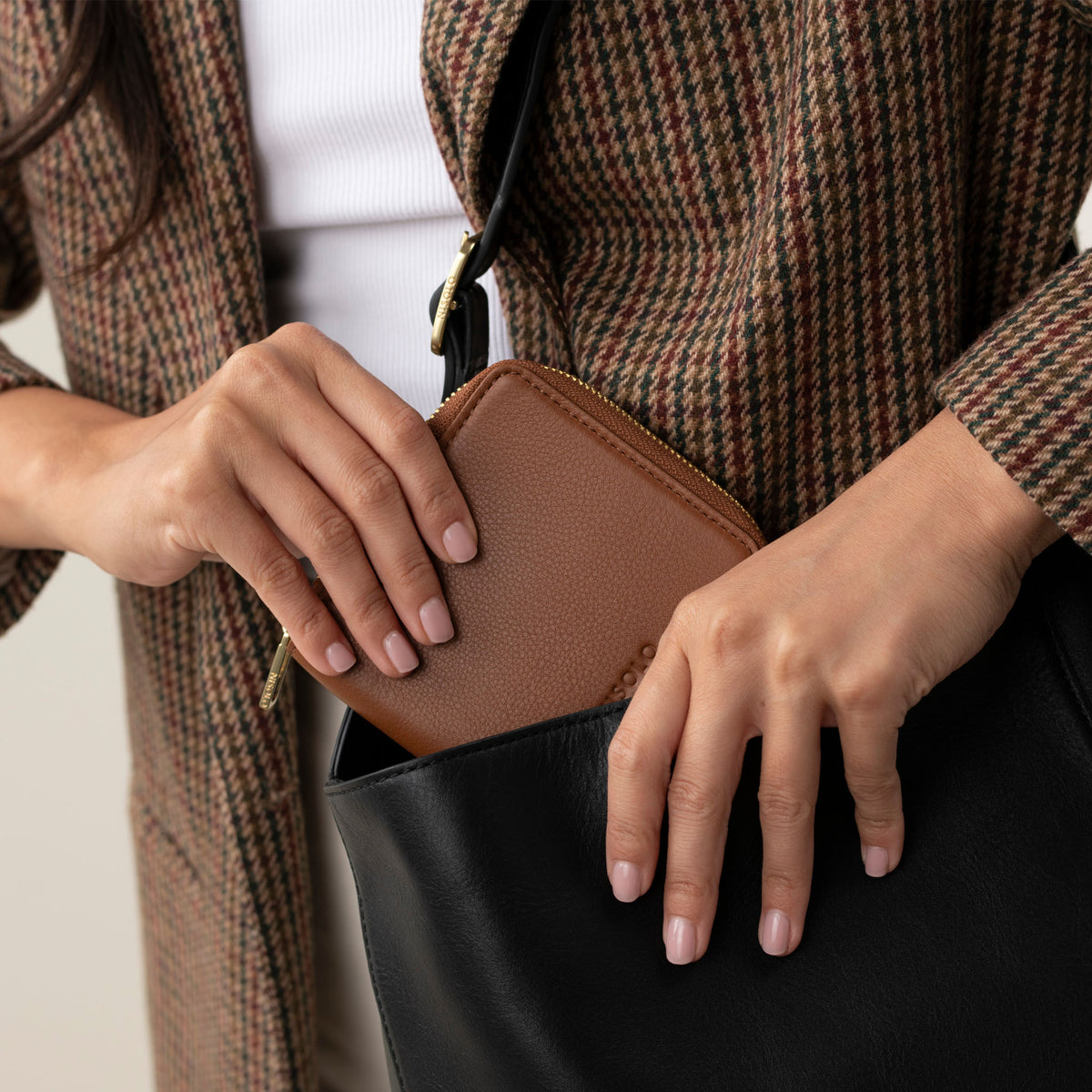 A person places a caramel brown rectangular zip wallet into a black bag. The wallet has a gold zipper and pebbled texture.