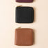 Three square leather NISOLO zip wallets in plum, black, and caramel arranged vertically on a beige background.