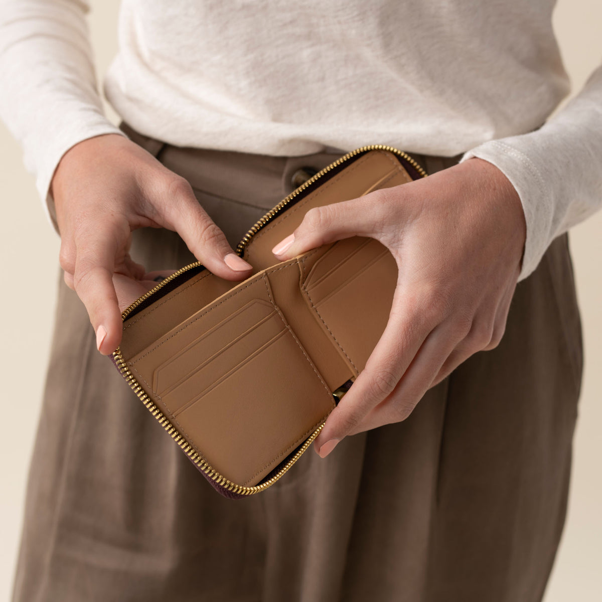 Hands holding an open, caramel-colored leather Remi Zip Wallet with card slots and a gold-toned zipper.
