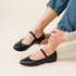 Pair of black leather Bea Go-To Mary Jane flats with elastic straps worn with blue denim jeans.