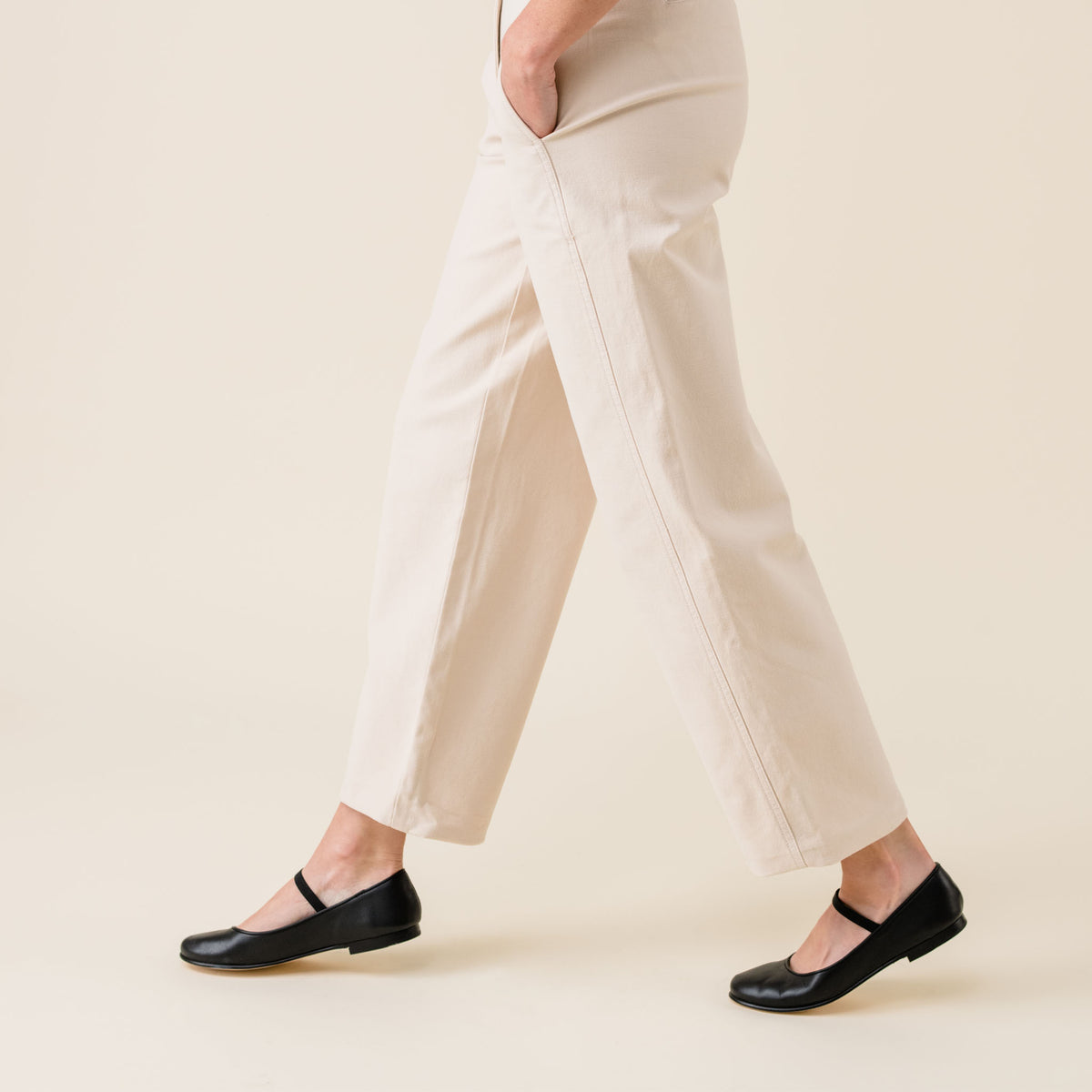 Pair of black leather Mary Jane flats worn by a person walking in tan trousers.