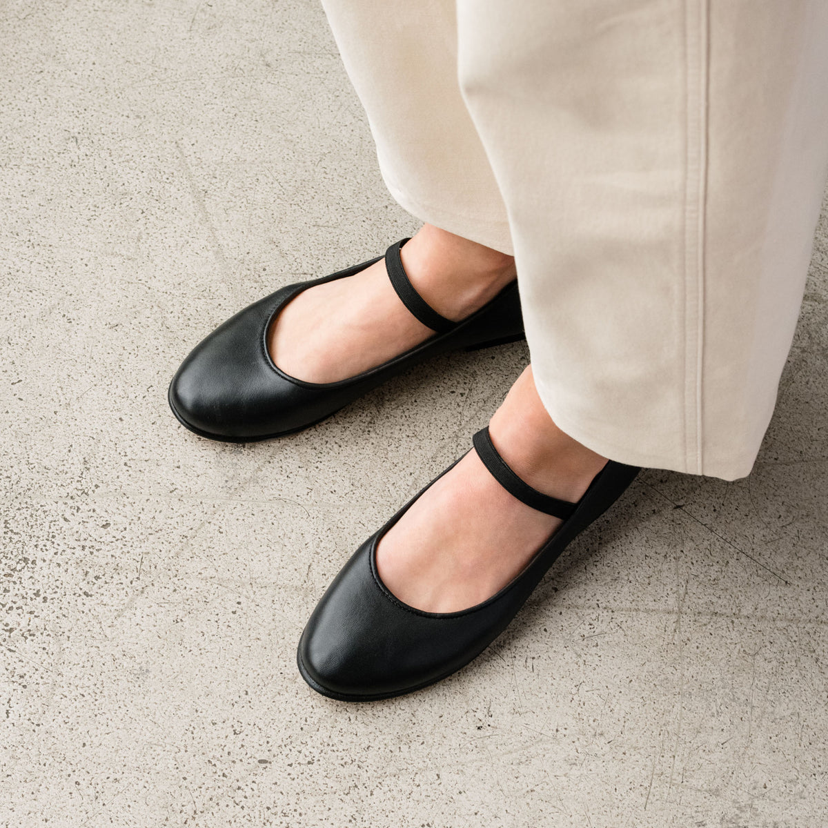 Pair of black leather Mary Jane flats with round toes and elastic straps worn with light-colored trousers.