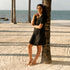 Woman in a black dress leaning against a palm tree on a beach wearing tan Catalina Slide Sandals in Almond.