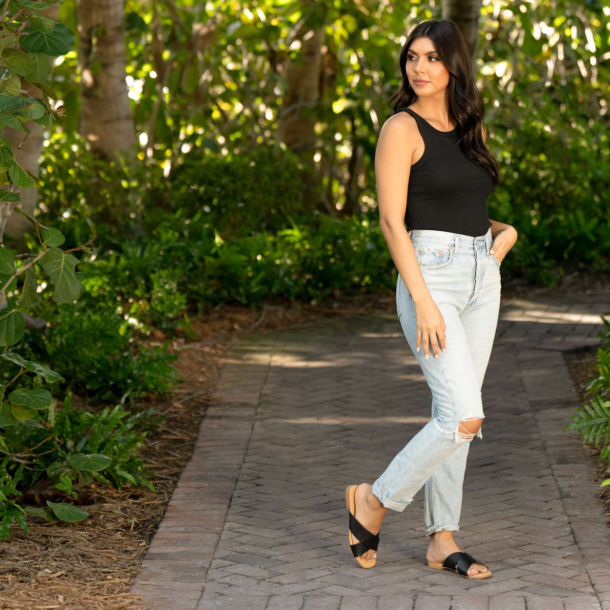 A woman wears black Catalina slide sandals with crossed straps on a stone path surrounded by green foliage.