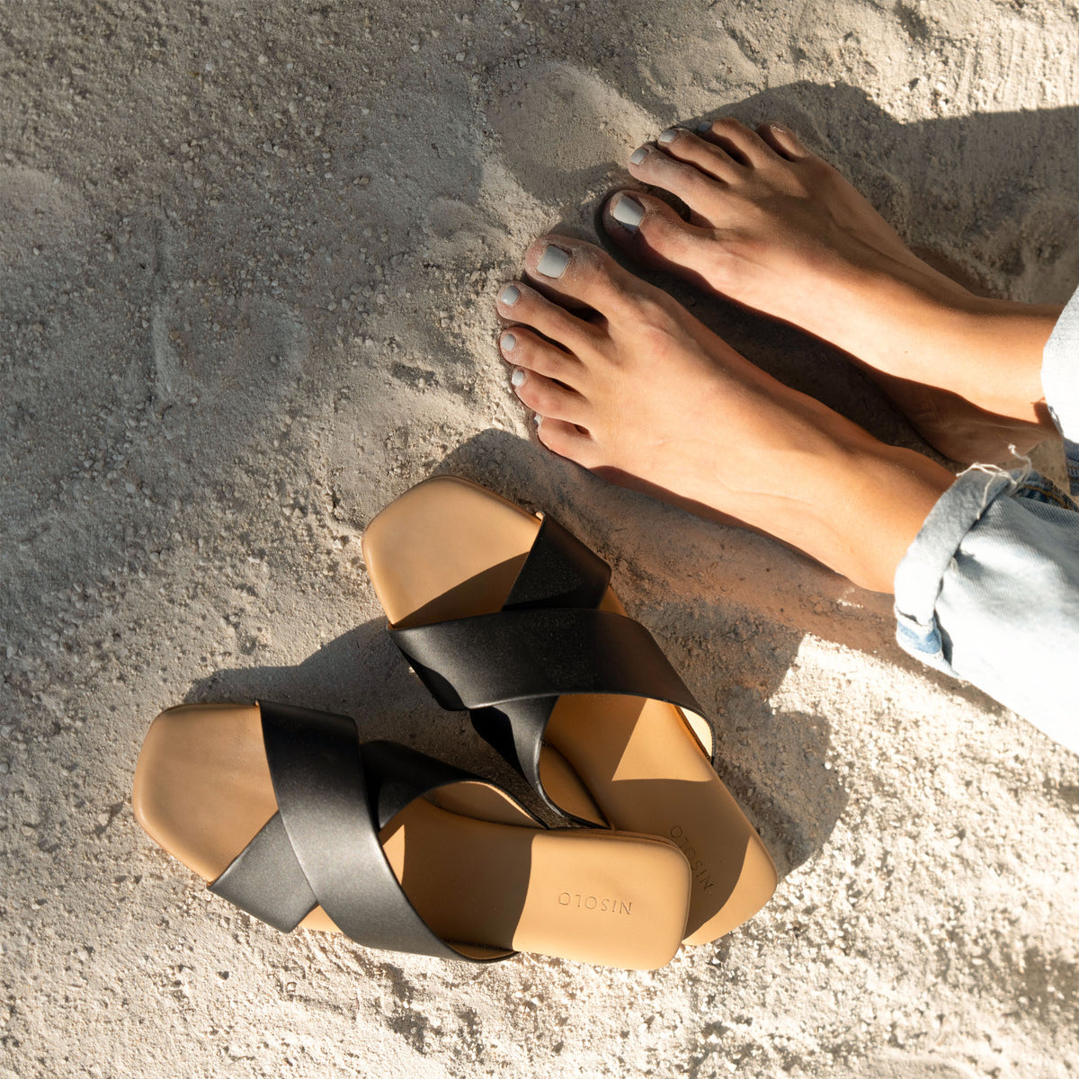 Two black leather Catalina Slide Sandals with tan footbeds by NISOLO resting on sand next to bare feet.