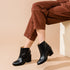 Pair of black leather ankle boots with block heels worn with brown trousers on a person sitting on a wooden box.