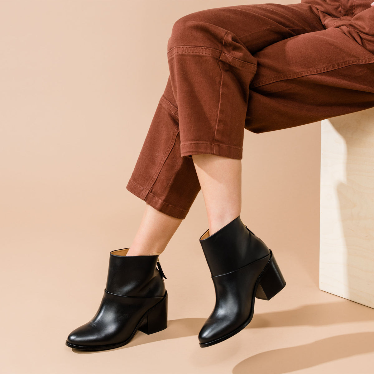 Pair of black leather ankle boots with block heels worn with brown trousers on a person sitting on a wooden box.