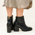 Pair of black leather ankle boots with block heels and gold-tone rear zippers, worn with a grey fabric skirt.