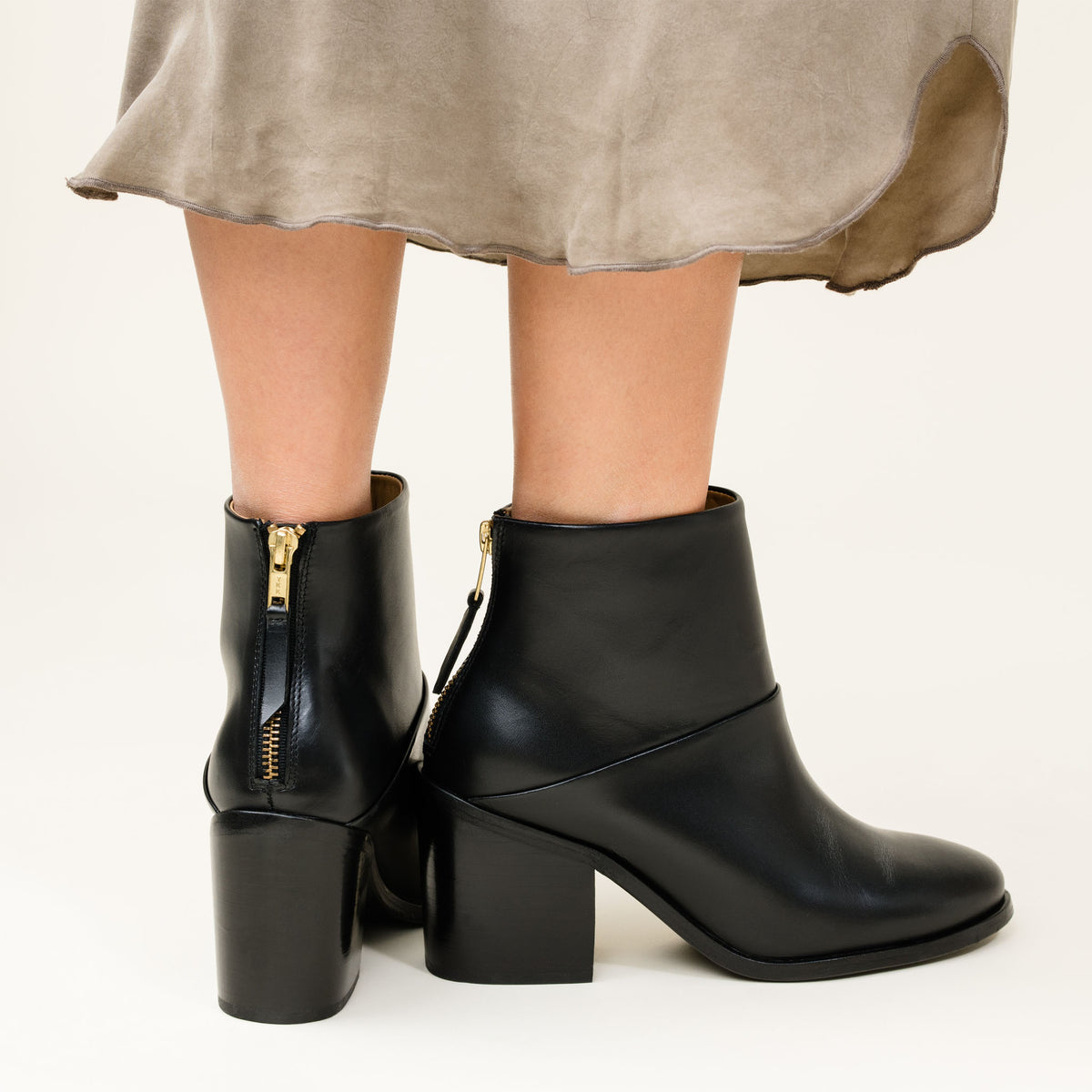 Pair of black leather ankle boots with block heels and gold-tone rear zippers, worn with a grey fabric skirt.