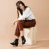 A woman sits on a wood box wearing black leather Dari Boot 2.0 ankle boots with a block heel and pointed toe.
