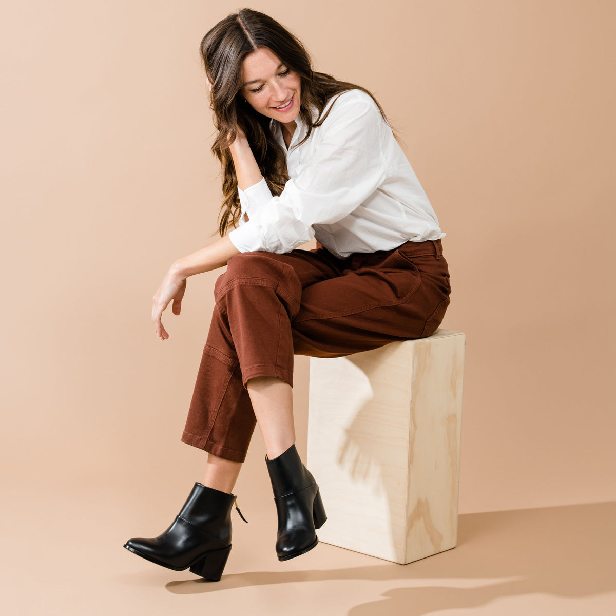A woman sits on a wood box wearing black leather Dari Boot 2.0 ankle boots with a block heel and pointed toe.