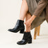 Pair of black leather ankle boots with block heels worn by a person sitting on a wooden stool.