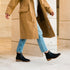 A person walking in black leather Eva Everyday Chelsea boots with tan soles, blue jeans, and a long tan coat.