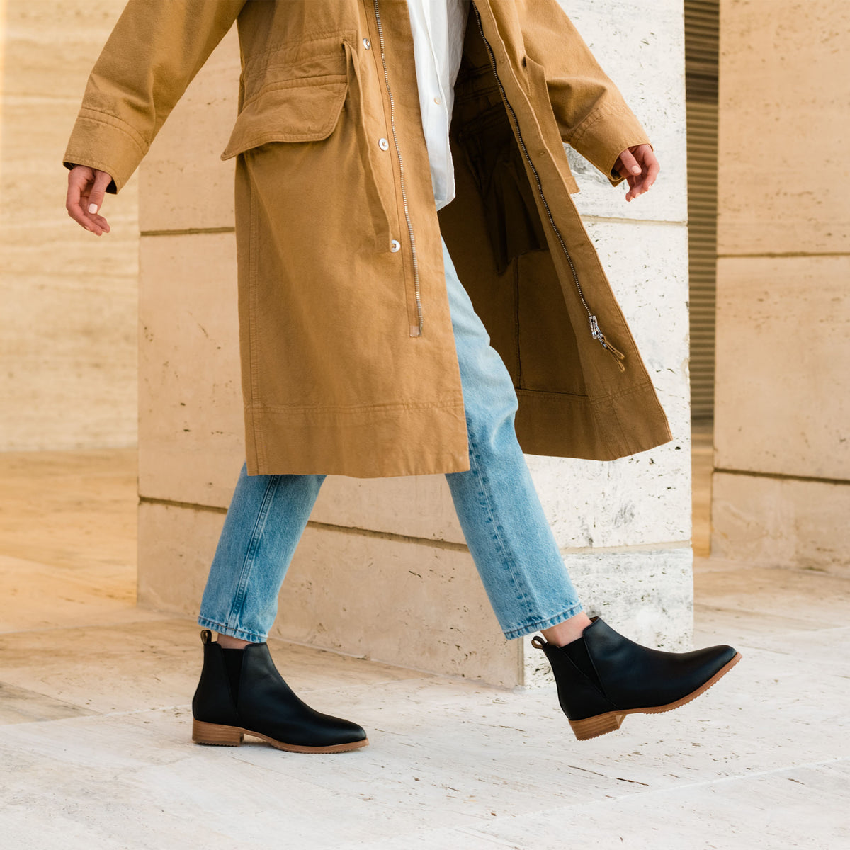 A person walking in black leather Eva Everyday Chelsea boots with tan soles, blue jeans, and a long tan coat.