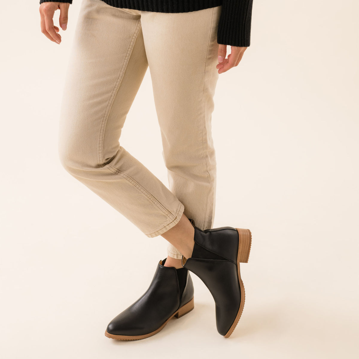 Pair of black leather Eva Everyday Chelsea Boots with tan soles worn with tan pants and a black sweater.