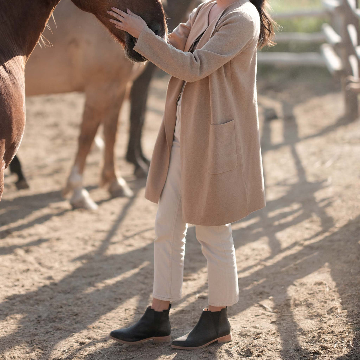 A person wearing black leather Eva Everyday Chelsea Boots, white pants, and a tan cardigan stands outdoors near a horse.