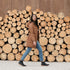A woman walks in black leather Chelsea boots, blue jeans, and a brown parka in front of a large stack of wood logs.