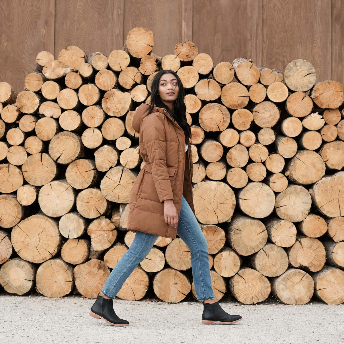 A woman walks in black leather Chelsea boots, blue jeans, and a brown parka in front of a large stack of wood logs.